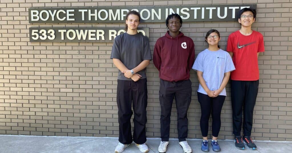 Boyce Thompson Institute students standing in front of brick wall with institute sign, diverse group of young researchers at a leading plant science and genomics research center.